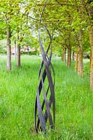 Flamme - sculpture en acier - dans l'herbe avec une allée d'arbres au-delà