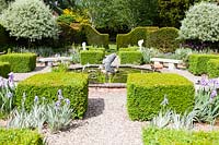 Vue sur l'étang de jardin avec les carrés topiaires environnants et les haies, plantation symétrique d'arbres, de plantes telles que l'iris dans le gravier et les sièges et les sculptures
