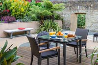 Salle à manger intime sur une terrasse en bois donnant sur une cour, bordée de parterres de fougères arborescentes australiennes, de cannas, de verveines et de sedums.