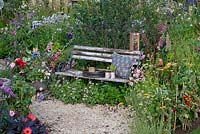 Jardin de cottage avec chemin de gravier serpentant à travers des parterres de fleurs et de légumes détendus. Un banc rustique se niche dans la plantation. BBC Springwatch Garden, conçu par Jo Thompson. RHS Hampton Court Palace Garden Festival, 2019.