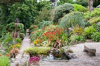 Un étang englouti dans le coin de la terrasse avec un chemin menant au-delà de parterres de fleurs luxuriants en pente. La plantation comprend des cordylines, des phormiums, Crocosmia 'Lucifer', des herbes et des euphorbes.