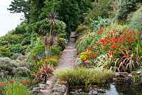 Un étang englouti dans le coin de la terrasse avec un chemin menant au-delà de parterres de fleurs luxuriants en pente. La plantation comprend des cordylines, des phormiums, Crocosmia 'Lucifer', des herbes et des euphorbes.