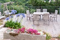 Terrasse avec mobilier de salle à manger encadrée de plantes succulentes dans une auge en pierre comprenant des sedums et des échéverias, entourée d'Erigeron karvinskianus autogerme et d'Agapanthus bleu.