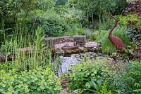 Un étang faunique bordé de joncs, ligularia, alchemilla et cranesbill, dominé par une terrasse en gravier avec des sièges en gabions.