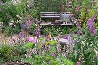 Outils de jardinage et coussin sur un banc de jardin rustique niché au milieu d'une plantation pérenne le long d'un chemin sinueux en gravier. RHS Hampton Court Festival 2019.