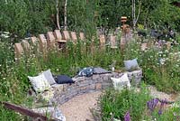 Coussin sur banc en pierre sèche incurvée soutenu par une palissade en bois, dans un jardin semi-sauvage. RHS Hampton Court Festival 2019.