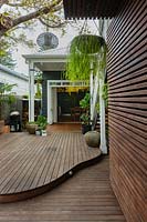 Un jardin montrant un platelage en bois à plusieurs niveaux avec un mur de garage en lattes de bois dur, un pot de homard recyclé, un Rhipsalis succulent dans un panier suspendu avec vue sur l'arrière d'une maison.