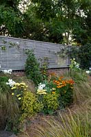 Vue du mur de briques de limite surmonté d'une clôture à lattes horizontales peinte en gris. Le parterre de fleurs devant le mur contient un obélisque et des plantes: Phlox 'David', Achillea 'Moonshine', Stipa arundinacea, Helenium 'Moerheim Beauty' et Alchemilla mollis