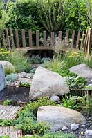 Piscine réfléchissante en acier corten avec pierres et rochers, banc incurvé avec Deschampsia cespitosa, Achillea 'Moonshine', Ophiopogon planiscapus 'Nigrescens' - Through Your Eyes Garden - RHS Hampton Court Palace Garden Festival 2019 - Conception: Lawrence Roberts et William Roobrouck Sponsors: Kebony, CED Stone, Produits métalliques R et G, William's Art and Design, Practicality Brown.