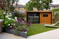 Jardin contemporain de Londres - vue sur le jardin de la maison vers la pelouse artificielle avec chaise longue, salle de jardin ou salle de gym et zone de terrasse en bois inférieure.