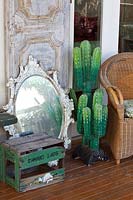 Une collection d'objets décoratifs sur une véranda, avec un cactus palster, une caisse de boisson gazeuse rétro, un miroir antique, une porte lambrissée et une chaise paon en rotin.