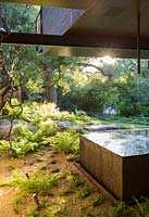 Pièce d'eau moderne en cuivre sur terrasse avec jardin de gravier sous une maison contemporaine, bois au-delà