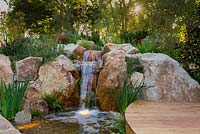 Une cascade construite à partir de gros rochers et de petits rochers, plantée d'herbes, de plantes à fleurs et de couvre-sols et dotée d'une promenade en bois courbé