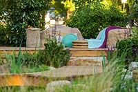 Une terrasse en bois avec des coussins, des jetés et un siège en jonc de mer, un environnement intercalé avec une variété d'arbustes, de plantes à fleurs et de feuilles à lanières