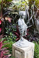 Feuillage tropical entourant la statue de chérubin sur socle, chérubin a chrysalide monarque accroché à une oreille