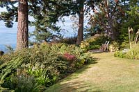 Voir la pelouse vers Puget Sound. Petit coin salon placé pour profiter de la vue. Parterres plantés avec une collection de plantes résistantes au cerf et résistantes à la sécheresse