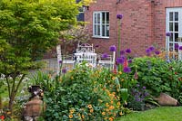 Un parterre de fleurs mélangé entre une pelouse et un coin salon sur la terrasse. Parterre de fleurs bombé planté de Geum 'Totally Tangerine', avens, alliums violets, nectaroscordum, lupins, géraniums et Acer palmatum 'Sango-kaku'