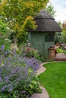 Une bordure de brique sépare la pelouse d'un parterre de fleurs herbacées de menthe des chats, de roses, d'ammi, de géraniums rustiques et d'alliums, menant à un pavillon d'été et à une terrasse en bois.
