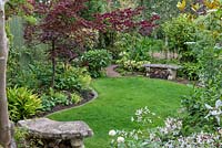 Une pelouse est entourée de parterres de fleurs incurvées de plantes aimant l'ombre telles que les fougères, le bistort, l'alchemilla, l'aucuba, les astilbes, les acers japonais et le Cercis canadensis 'Forest Pansy ''.