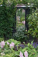 Au-dessus d'une touffe de Gillenia trifoliata, un miroir est fixé à une clôture limite, reflétant les astilbes roses et les ligularias jaunes au premier plan.