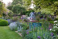 Un parterre de gravier est planté d'épis d'agneau argenté, d'alliums, d'iris et de centranthus, entourant un grand tambour métallique de dahlias rouges et de lierre. Au-delà, un pavillon d'été donne sur une terrasse et un parterre de fleurs herbacés ponctués d'obélisques roses.