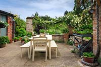 La terrasse abritée avec table et chaises peintes en crème, et Rosa 'Alberic Barbier' entraînée le long de butées de corde épaisse