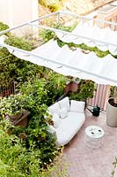 Petite terrasse carrelée vue de la maison, montrant un auvent ombragé sur un coin salon soutenu par des plantes en pot