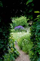 Vue à travers l'arc découpé dans une haie avec un chat sur un chemin de gravier. Pelouse et parterres de jardin de chalet