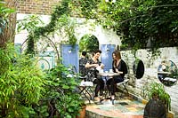 Deux femmes assises prenant le thé sur le patio carrelé, dans un espace clos de murs de briques peintes avec miroirs et plantes