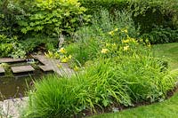 Un jardin de campagne de banlieue informel avec des plantes vivaces, comme Hemerocallis, dans un parterre de fleurs près d'un petit étang avec des pavés et des tremplins