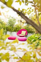 Espace détente sur une terrasse vue à travers les arbustes. Paravents mixtes chaise et coussins rose vif