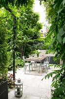 Vue à travers le feuillage de la terrasse pavée blanche avec coin repas et cuisine extérieure, les bâtiments voisins grillagés par des clôtures métalliques et des plantes à feuillage