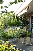 Disposition de bacs plantés sur une terrasse pour diviser la zone en zones de détente et de restauration. Les jardinières contiennent un feuillage mixte d'herbes ornementales, de vivaces et d'arbustes. Auvent d'ombre attaché au plafond.