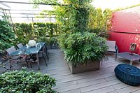 Vue d'ensemble du jardin sur le toit en bois avec salle à manger sous pergola, une cloison rouge et des jardinières avec des arbustes pour le dépistage