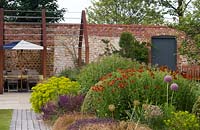 Pergola en acier corten au-dessus du coin salon contre le mur de briques d'un jardin de campagne clos, dans le coin au premier plan d'un parterre de fleurs vivaces