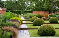 Jardin de campagne moderne et formel avec chemin aménagé, pelouse divisée par des bordures en corten avec des dômes topiaires et des parterres de fleurs vivaces