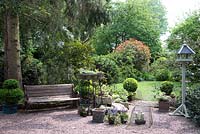 Vue sur un jardin ombragé avec gravier, banc, pots, topiaire avec pelouse, arbres et arbustes