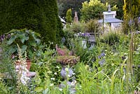 Jardin clos en juin plein de plantations luxuriantes, de pots et de structures, dont une maison de thé sur pilotis