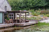 Une terrasse en bois de bois amène la maison en contact étroit avec le grand étang qui occupe le site d'une ancienne unité de porcs intensifs à Am Brook Meadow, Devon en août.