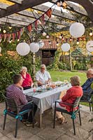 Les gens à table à manger en appréciant les snakcs et les boissons sous une pergola en bois décorée de chaînes légères et de banderoles colorées