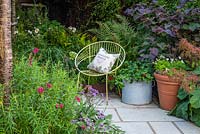 Une chaise moderne et un pot en terre cuite d'alto blancs, et un récipient agricole en métal galvanisé planté de pélargoniums. Derrière, un parterre surélevé planté d'anémone, d'herbes, de fougères et de Cercis canadensis.