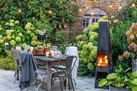 Un coin repas dans une cour-jardin éclairée par une guirlande d'ampoules et de bougies, chauffée par une cheminée extérieure moderne. La plantation derrière comprend l'Hortensia 'Limelight', Rosa 'Blush Noisette', Anemone hybrida 'Elfin Swan' et des fougères.