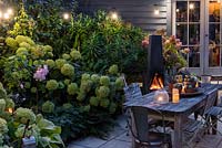 Une salle à manger dans une cour-jardin éclairée par une guirlande d'ampoules et de bougies, chauffée par une cheminée extérieure moderne. La plantation derrière comprend des hortensias 'Limelight', des rosiers 'Blush Noisette' et des fougères.