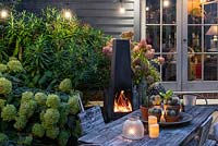 Un coin repas dans une cour-jardin éclairée par une guirlande d'ampoules et de bougies, chauffée par une chiminea extérieure moderne. La plantation derrière comprend l'Hortensia 'Limelight', la Rosa 'Blush Noisette' et les fougères.