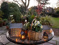 Un affichage de table de plantes d'hiver, y compris Helleborus, Ilex, Cotoneaster et Ilex éclairé par des bougeoirs en forme de maison dans un jardin au crépuscule.