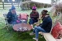 Des femmes assises autour d'un feu dans un foyer en acier corten, buvant du vin chaud un jour d'hiver glacial.