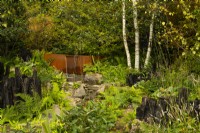 Une forêt contenant une fontaine et un ruisseau coulant d'une auge en acier sur des rochers entourés d'arbres, de vivaces et de parterres de rondins carbonisés dans le jardin biologique de la vallée de Yeo.