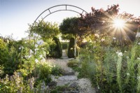 Jardin de gravier en juillet avec plantes herbacées et conifères taillés encadrés d'une arche métallique.
