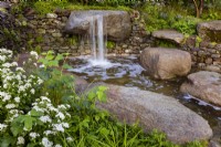 Jardin du Psaume 23. Cascade naturaliste d'eau de granit, roches usées dans un mur de pierres sèches dans une piscine tranquille. Avec Eupatorium 'Lucky Melody' et de jeunes pousses de Viburnum opulus .