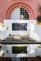 Le lac serpent avec vue sur la maison et panneau décoratif de ciment peint dans le mur au-dessus des marches, reflété dans la piscine.
